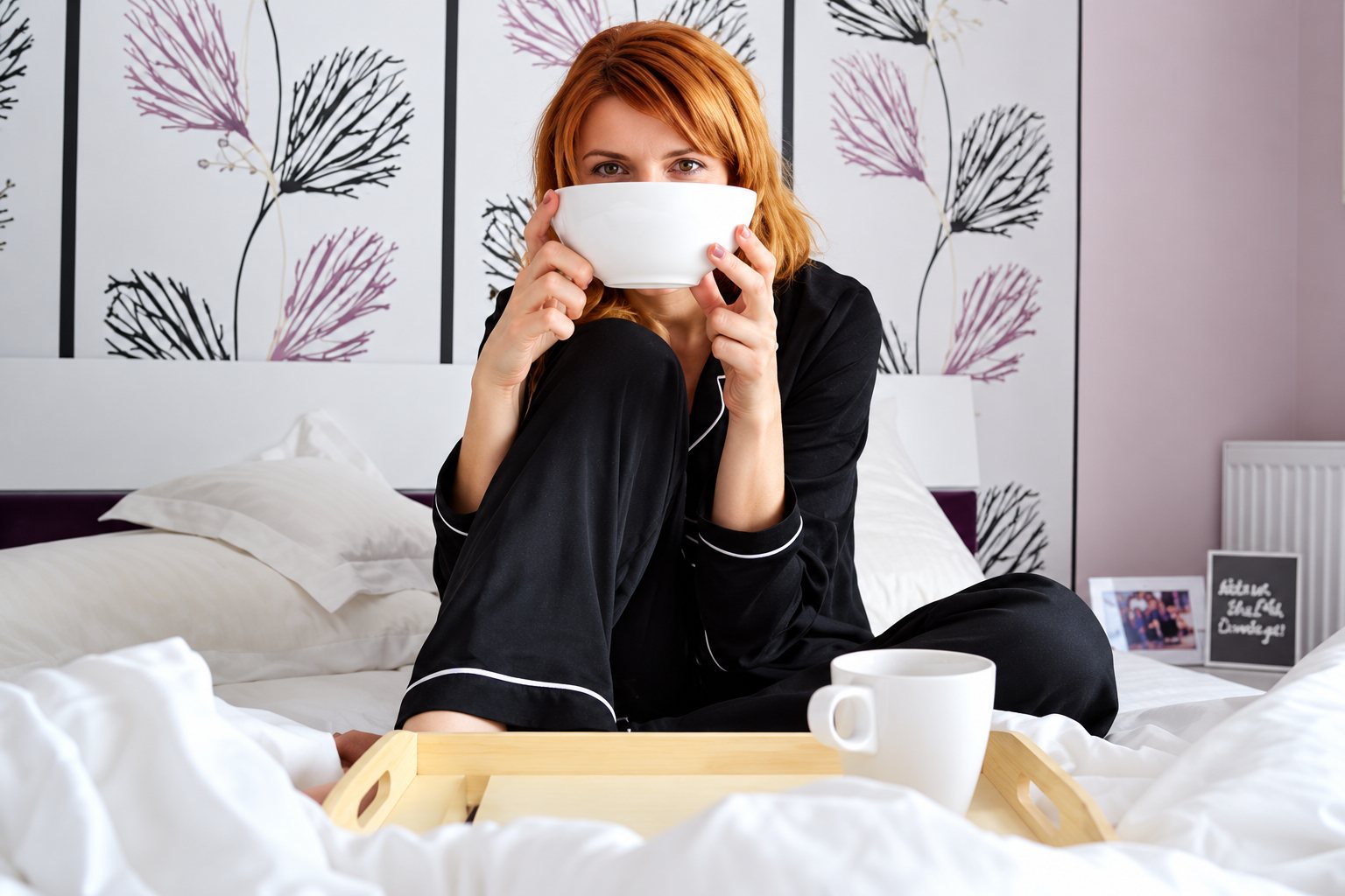 Une femme aux cheveux roux, en pyjama noir, est assise sur un lit défait et tient un grand bol blanc devant son visage, avec un plateau et une tasse devant elle.