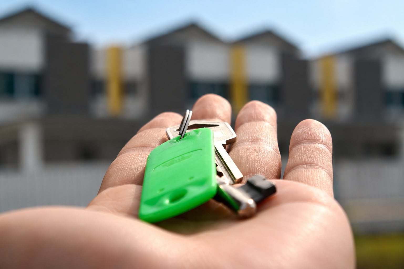 Une main ouverte présente un trousseau de clés avec un porte-clés vert, devant des maisons modernes floues en arrière-plan.